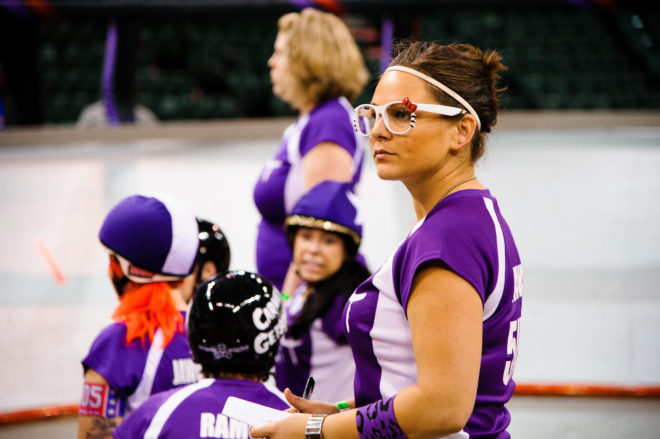 Image of Anayalator in a purple jersey and white Hello Kitty glasses watching the banked track with roller derby skaters in the background